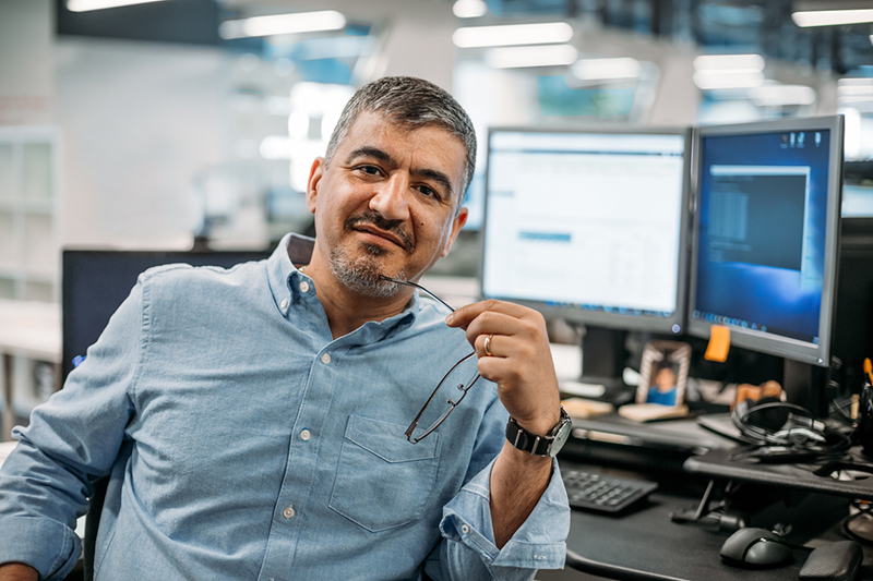 A man in a blue shirt holding glasses, sitting at a desk with computer monitors.